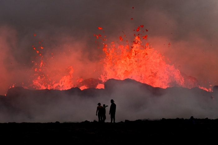 People watch flowing lava during a volcanic eruption near Litli Hrutur, south-west of Reykjavik in Iceland on July 10, 2023. - A volcanic eruption started on July 10, 2023 around 30 kilometers (19 miles) from Iceland's capital Reykjavik, the country's meteorological office said, marking the third time in two years that lava has gushed out in the area. "The eruption is taking place in a small depression just north of Litli Hrutur, from which smoke is escaping in a north-westerly direction," the office said.  Footage circulating in the local media shows a massive cloud of smoke rising from the ground as well as a substantial flow of lava.  (Photo by Kristinn Magnusson / AFP) / Iceland OUT / ICELAND OUT
